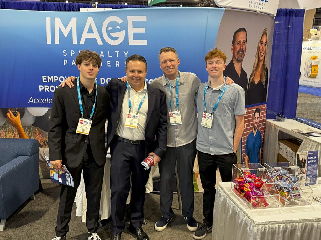 Four people posing and smiling at a trade show booth for Image Specialty Partners. The backdrop features the company logo and promotional graphics. They are wearing conference badges and standing beside a display table with giveaways.